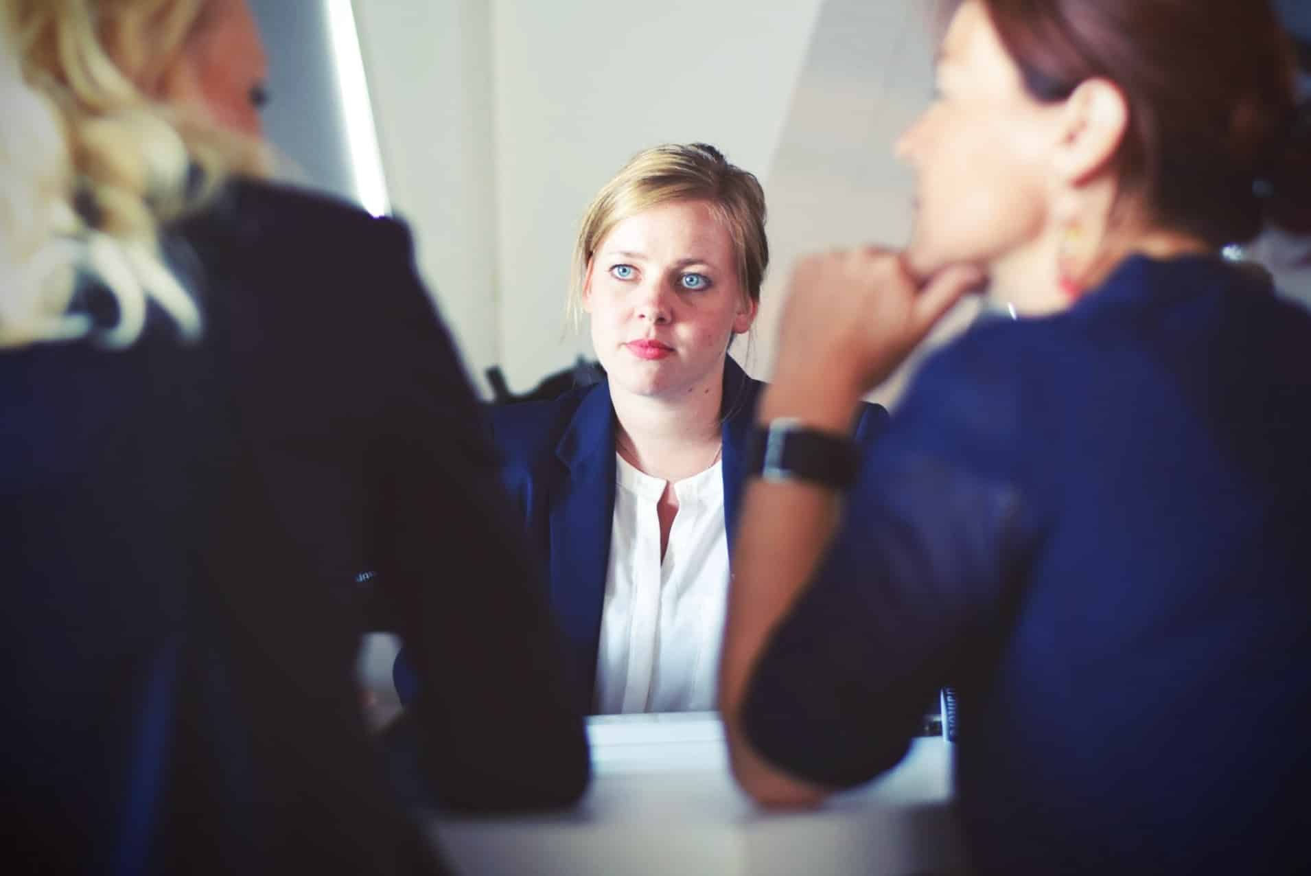A blond female professional sits in a meeting facing two other women.